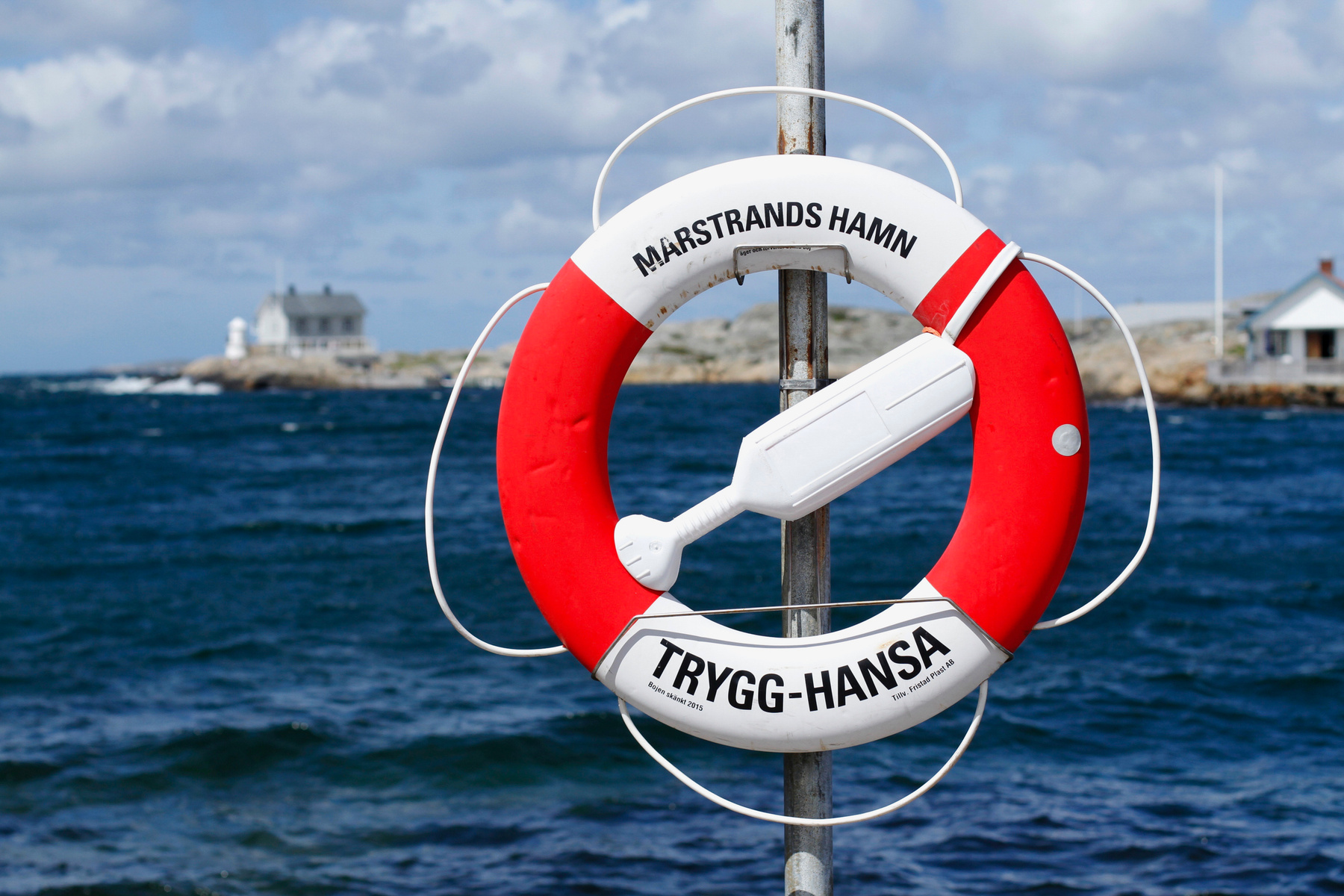 En röd och vit livboj med texten "MARSTRANDS HAMN" och "TRYGG-HANSA" är monterad på en metallstolpe. Bakgrunden visar ett blått hav under en molnig himmel, med en byggnad och en fyr synliga i fjärran. Bilden är tagen utomhus.