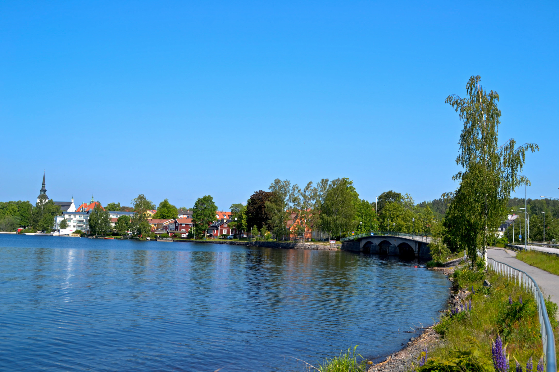 En vidvinkelbild som visar en sjö med en stad och en kyrka i bakgrunden. Ett bro går över sjön. Till höger finns en gång- och cykelväg med ett staket och grönska. Himlen är klarblå. Till vänster i sjön speglar husen och träden sig i vattnet.