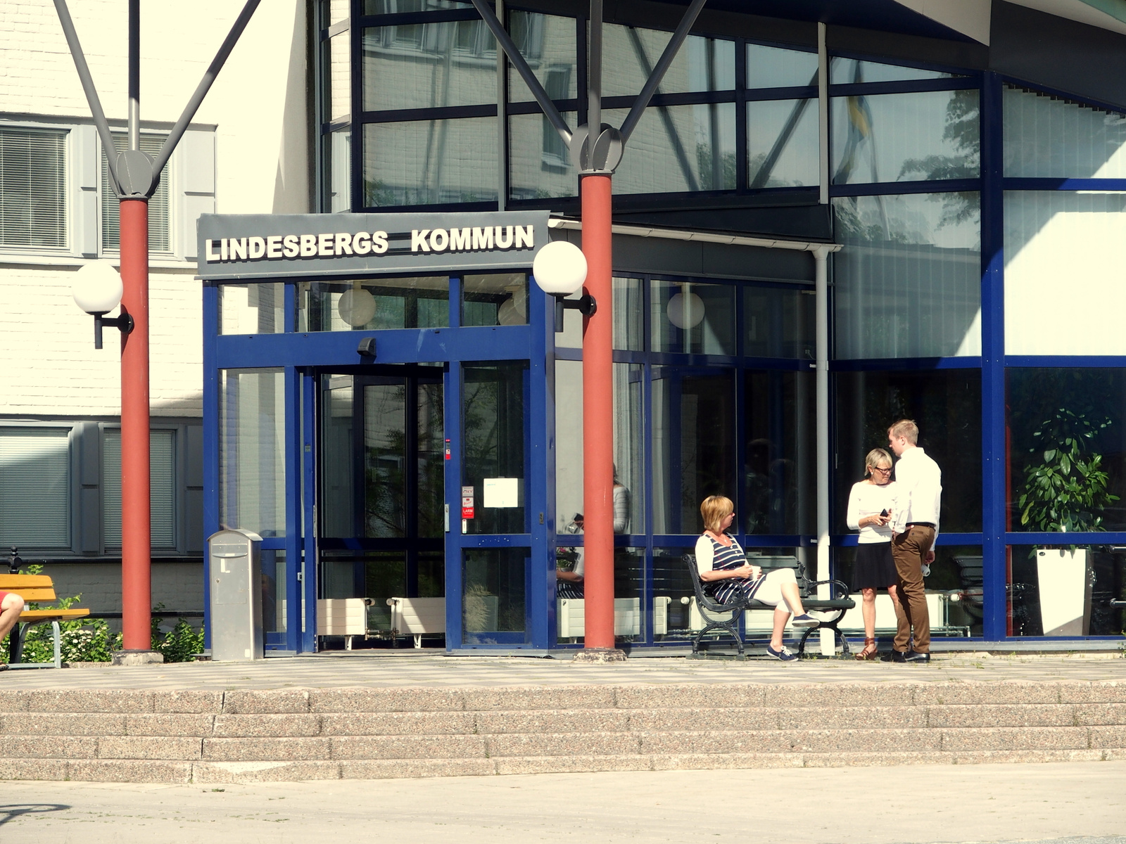 En fasadbild av Lindesbergs kommunhus en solig dag. Tre personer syns utanför entrén. En kvinna sitter på en bänk, medan en man och en kvinna står och pratar. Byggnaden har stora fönster och en blå entré, med en skylt där det står "Lindesbergs kommun