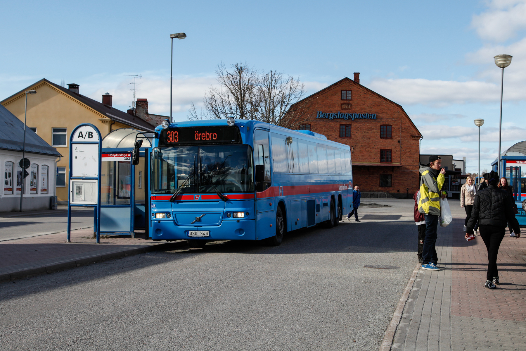 En blå buss med "303 Örebro" syns i bildens centrum, stående vid en hållplats. Flera personer befinner sig på trottoaren intill bussen, och i bakgrunden syns tegelbyggnader under en klarblå himmel med moln.