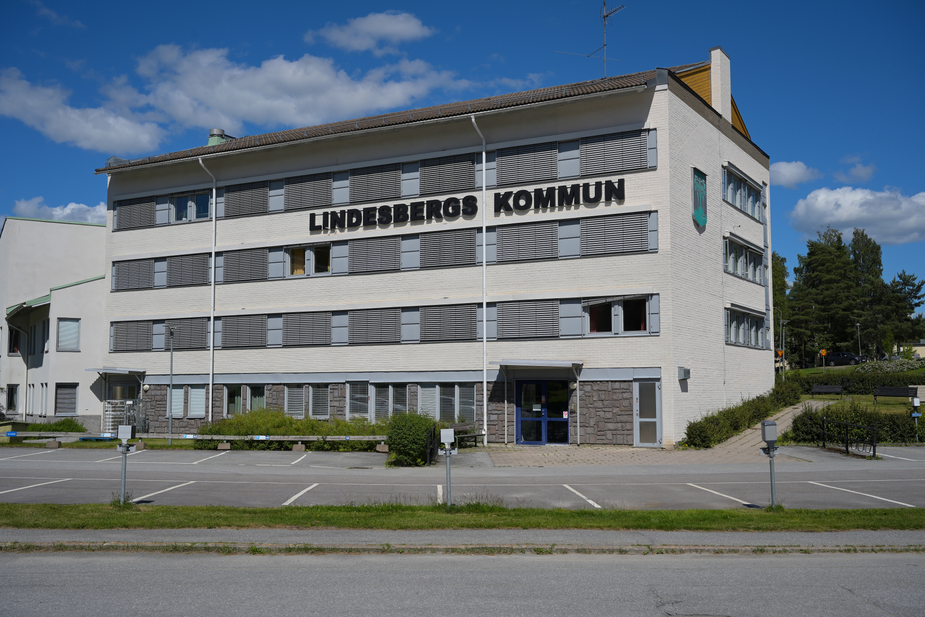 En vit tegelbyggnad med texten "LINDESBERGS KOMMUN" på fasaden, omgiven av en parkeringsplats med vita linjer, grönska, och träd under en blå himmel med vita moln.