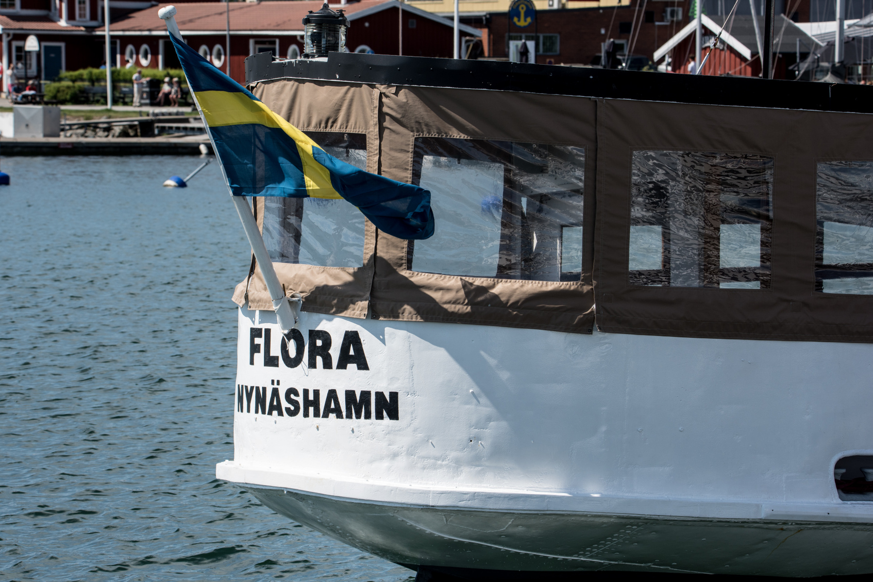 En vit båt med texten "FLORA NYNÄSHAMN" på aktern ligger i vattnet. En svensk flagga vajar från aktern. I bakgrunden syns röda byggnader och bryggor med människor.