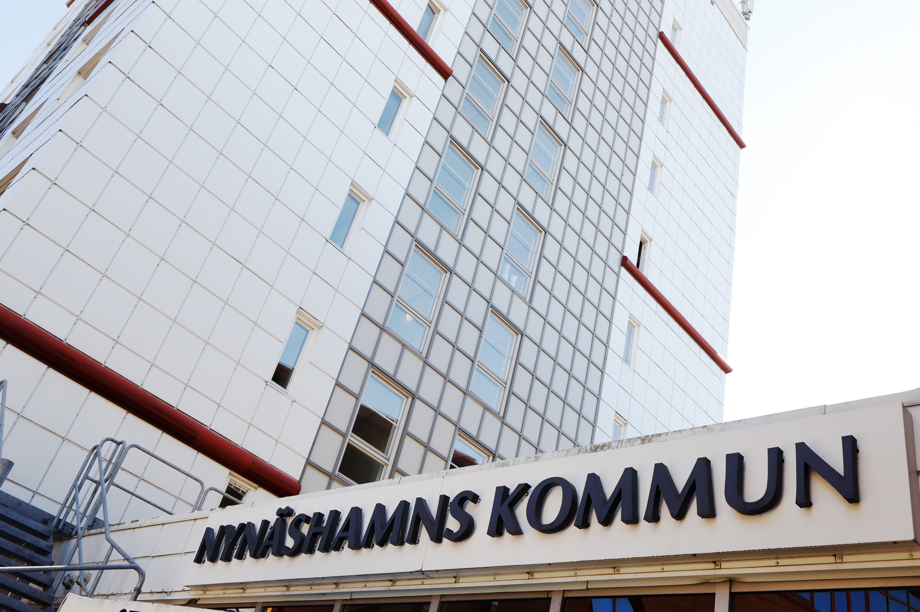 En modern byggnad med en fasad av vita plattor och grå glaspaneler, sett underifrån. Ovanför entrén finns en skylt med texten ”NYNÄSHAMNS KOMMUN”. Byggnaden har horisontella röda bandpartier och flera fönster. Himmelen är ljus och syns i bakgrunden.