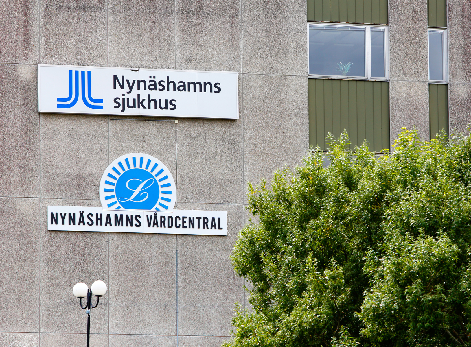 En bild som visar en del av en byggnadsfasad med skyltar för "Nynäshamns sjukhus" och "Nynäshamns vårdcentral". Skyltarna är vita med blå text och logotyper. Till höger om byggnaden står ett grönskande träd, och i förgrunden syns en gatlampa.