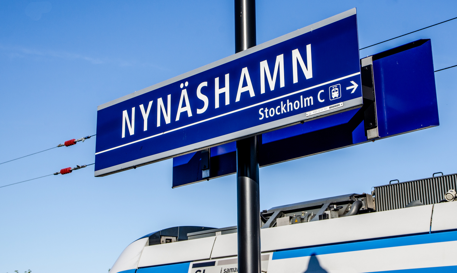 En blå skylt med texten "NYNÄSHAMN" och "Stockholm C" samt en riktningspil och tågsymbol, monterad på en svart stolpe. En del av ett vitt och blått tåg är synligt i bildens nedre del. Himlen är klarblå och ljus.
