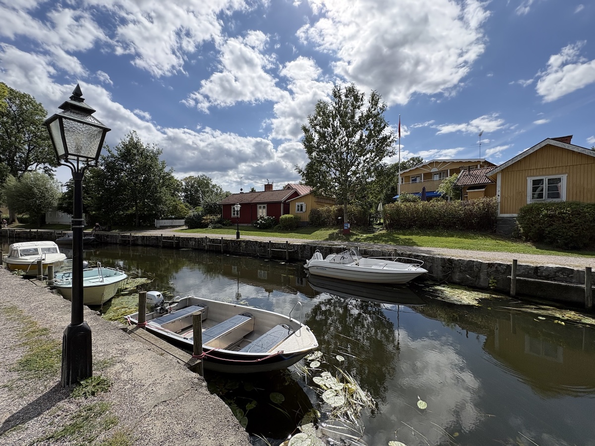 En bild som visar en smal kanal med båtar förtöjda längs sidorna, varav en lyktstolpe står i förgrunden till vänster. Bakom kanalen syns flera hus i olika färger, samt träd och buskar under en blå himmel med moln.