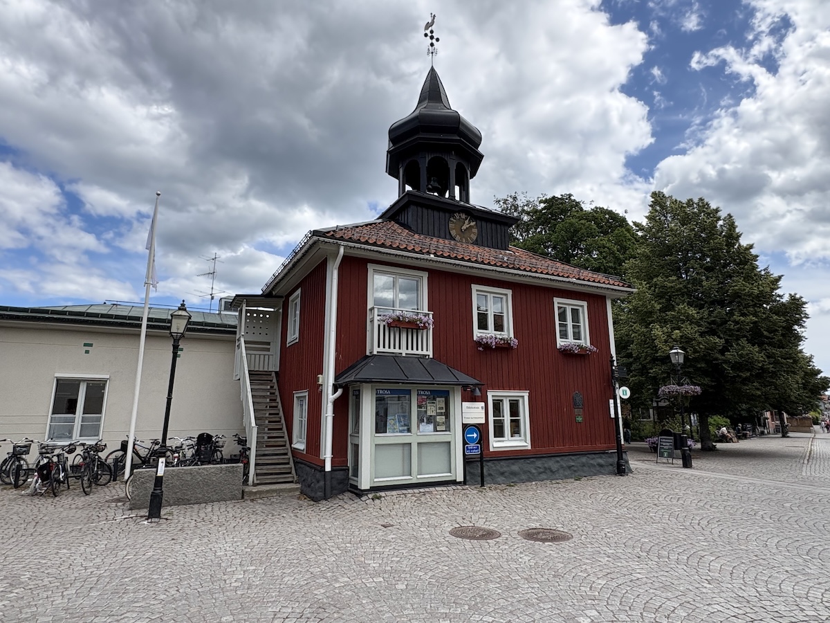 En röd träbyggnad med vit spröjsade fönster och en svart takspira med en vindflöjel. Till vänster om byggnaden syns en trappa som leder upp till en dörr och längre bort en ljusare byggnad. Ett antal cyklar är parkerade vid den ljusare byggnaden. Fram