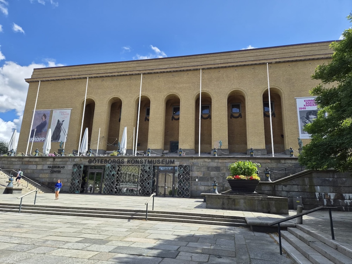 En gul tegelbyggnad med valvformade fönster och pelare under en blå himmel med vita moln. Framför byggnaden finns trappor, en plantering och skyltar med texten "GÖTEBORGS KONSTMUSEUM" och "HASSELBLAD CENTER". Flera människor syns gå på trapporna och