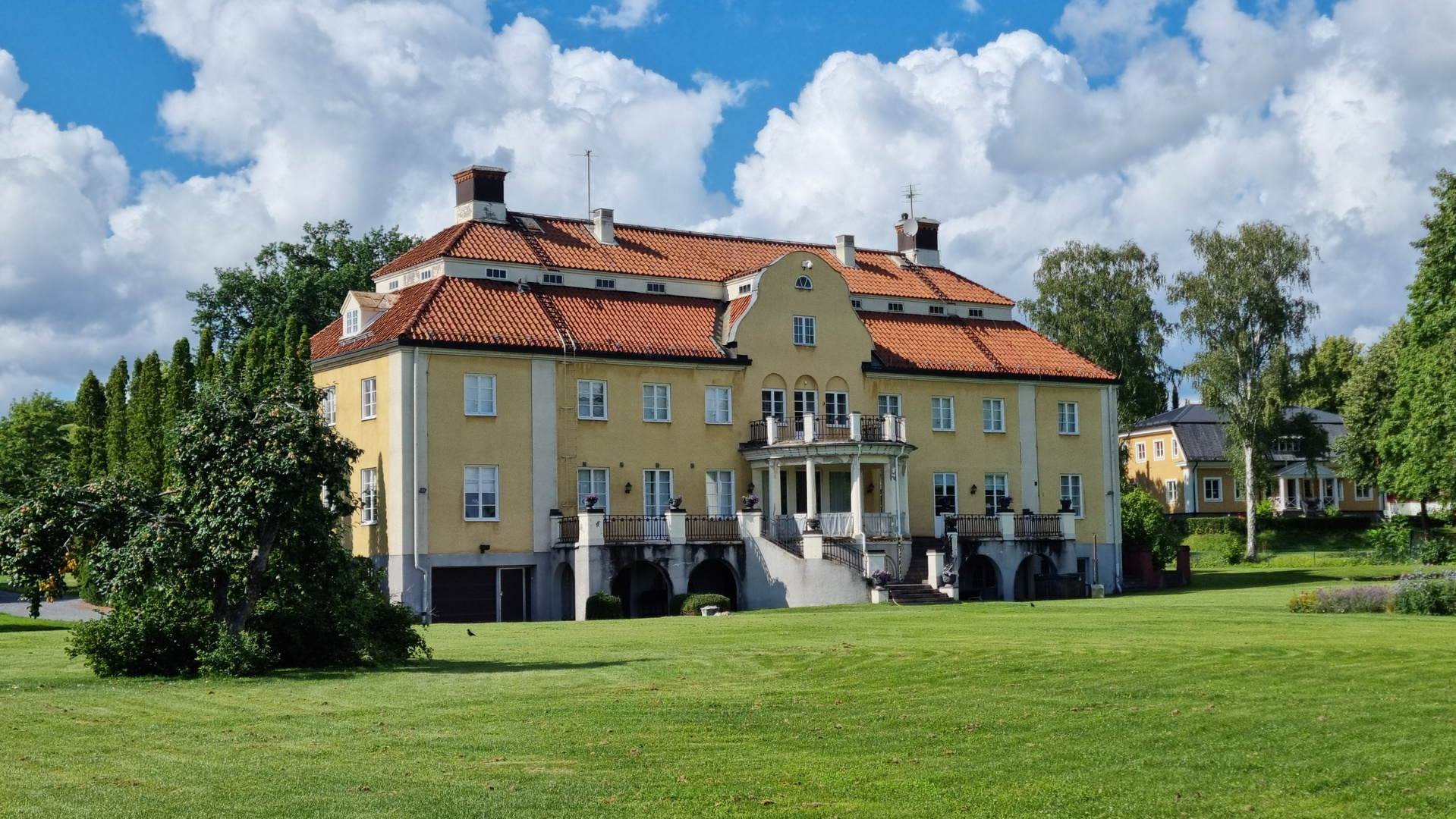 En stor, gul byggnad med rött tegeltak, flera fönster och en balkong med pelare, sedd från en gräsyta. Byggnaden har en stor entrétrappa och omges av träd och blå himmel med vita moln.