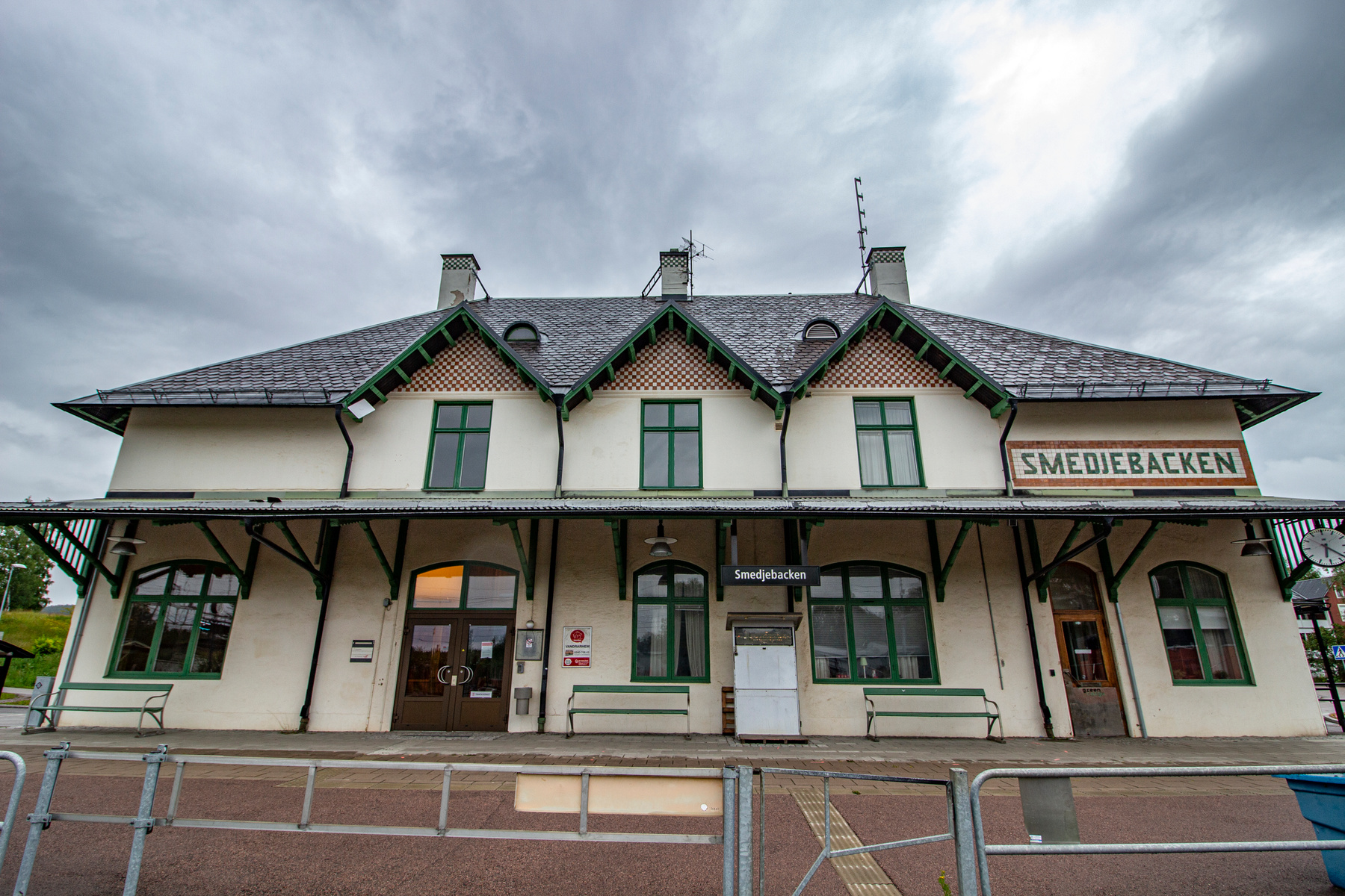 En stor, ljusbeige byggnad med mörkgröna fönsterramar och takdetaljer, samt ett tak av mörkt skiffer eller plåt. Byggnaden har flera skorstenar och takkupor. En skylt med texten "Smedjebacken" syns på byggnaden och även under takutsprånget. Framför b