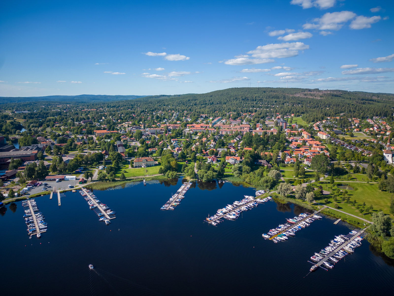 En vidvinkelbild tagen uppifrån över en sjö med ett flertal båtar förtöjda vid bryggor. Längs sjöns strandlinje syns en tätbebyggd stad med bostadshus, grönområden och skogklädda höjder i bakgrunden under en klarblå himmel med några moln.
