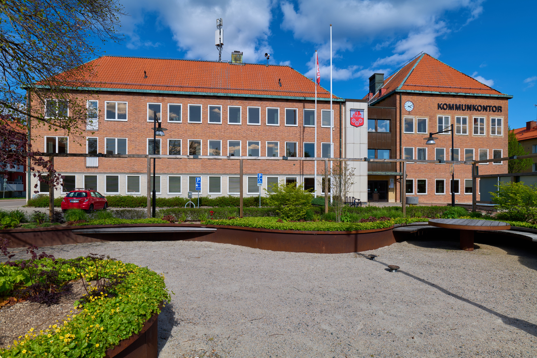 En röd tegelbyggnad med texten "KOMMUNKONTOR" och en klocka på fasaden. Framför byggnaden finns ett grönområde med planteringar, grusade ytor och sittplatser. En röd bil är parkerad vid sidan av byggnaden.