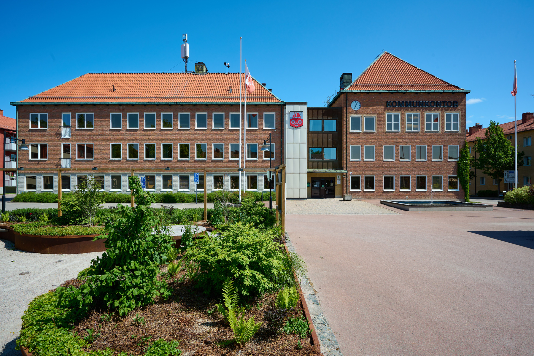En stor, röd tegelbyggnad med tegeltak och många fönster dominerar bilden under en klarblå himmel. Framför byggnaden finns ett öppet torg med en fontän, en flaggstång med en flagga, samt planteringar med gröna buskar och växter. På byggnadens fasad s