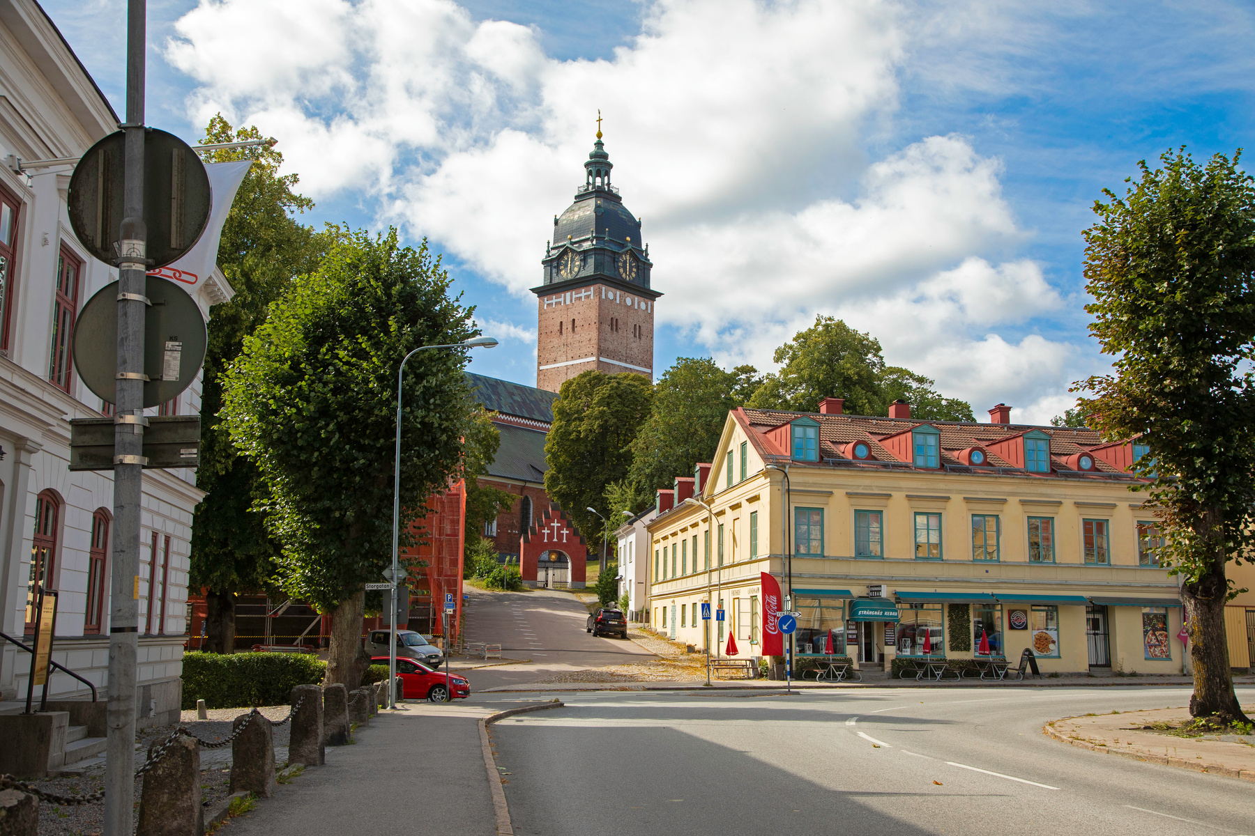 En gatuscen från Strängnäs med Strängnäs domkyrka i bakgrunden. Ett gult hus med restauranger på bottenvåningen syns till höger. På trottoaren syns flera träd och lyktstolpar. En röd bil är parkerad vid trottoarkanten och moln syns på himlen.