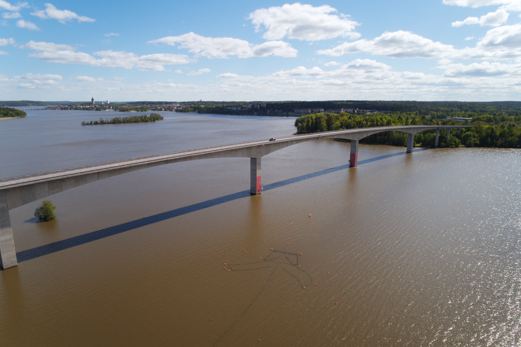 En vidvinkelbild från ovan visar en lång bro som sträcker sig över en sjö. På bron kör en mörk bil. Sjön omges av gröna träd och i bakgrunden syns en stadssiluett. Under bron syns en kedja av bojar i vattnet.