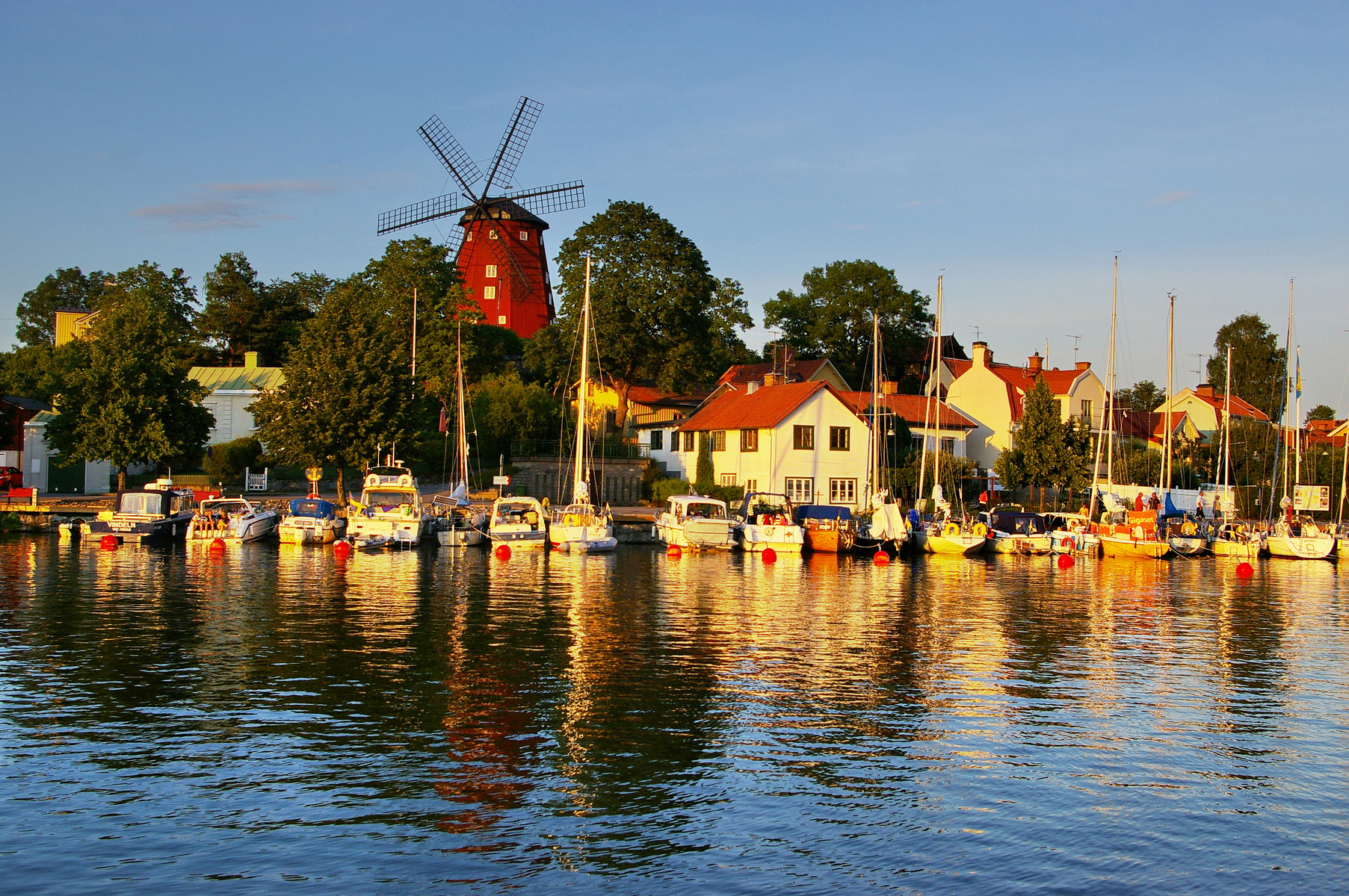 En bild som visar en småbåtshamn med flera segelbåtar och motorbåtar förtöjda längs en strandkant. Bakom båtarna syns hus med röda tegeltak och grönskande träd. I bakgrunden reser sig en röd väderkvarn. Vattenytan speglar himlen och de omgivande bygg