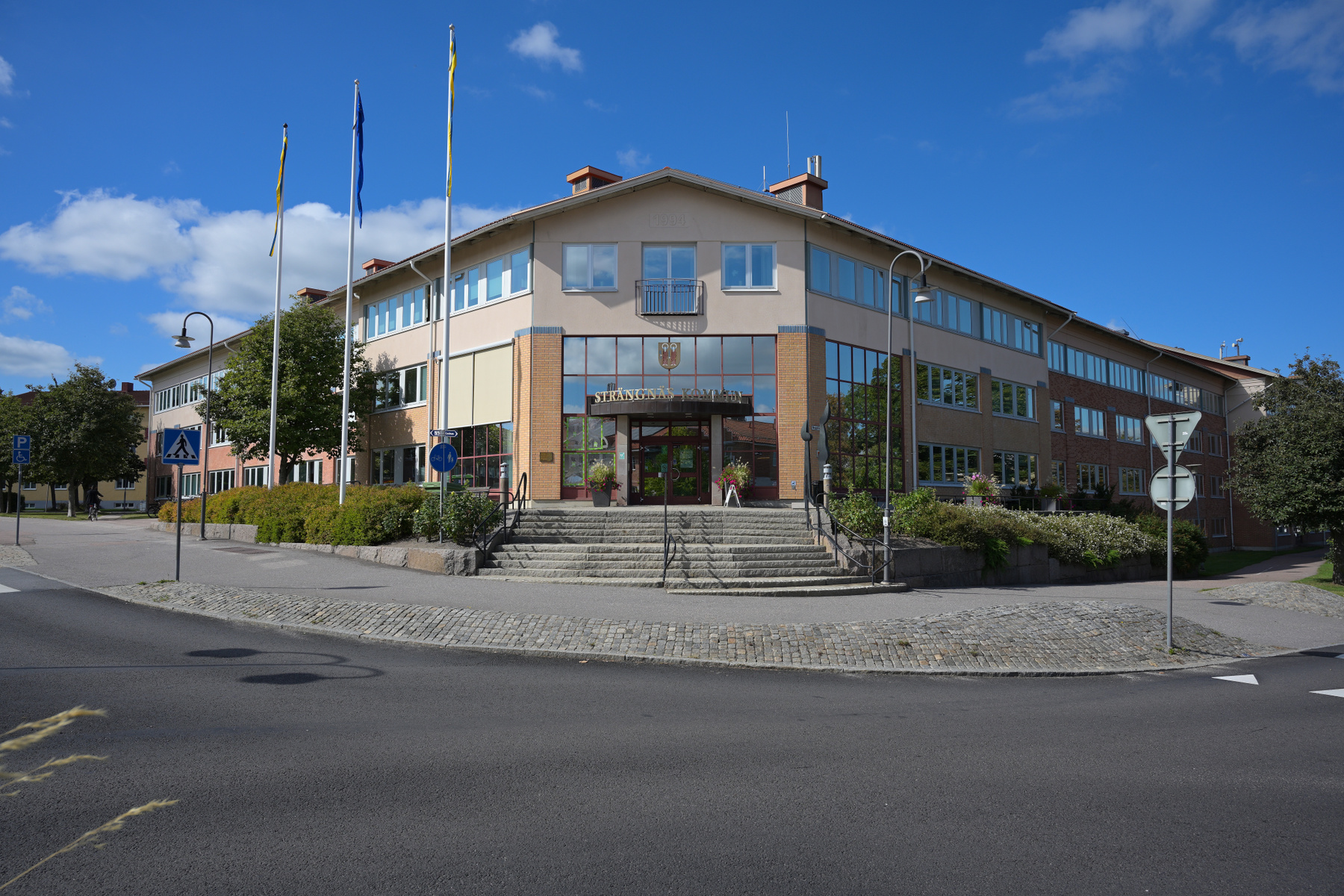 En stor byggnad med flera våningar och en framträdande ingång med trappor dominerar bilden. Byggnaden är omgiven av grönska, flaggstänger och en asfalterad väg med trottoarer. En blå himmel med vita moln är synlig.