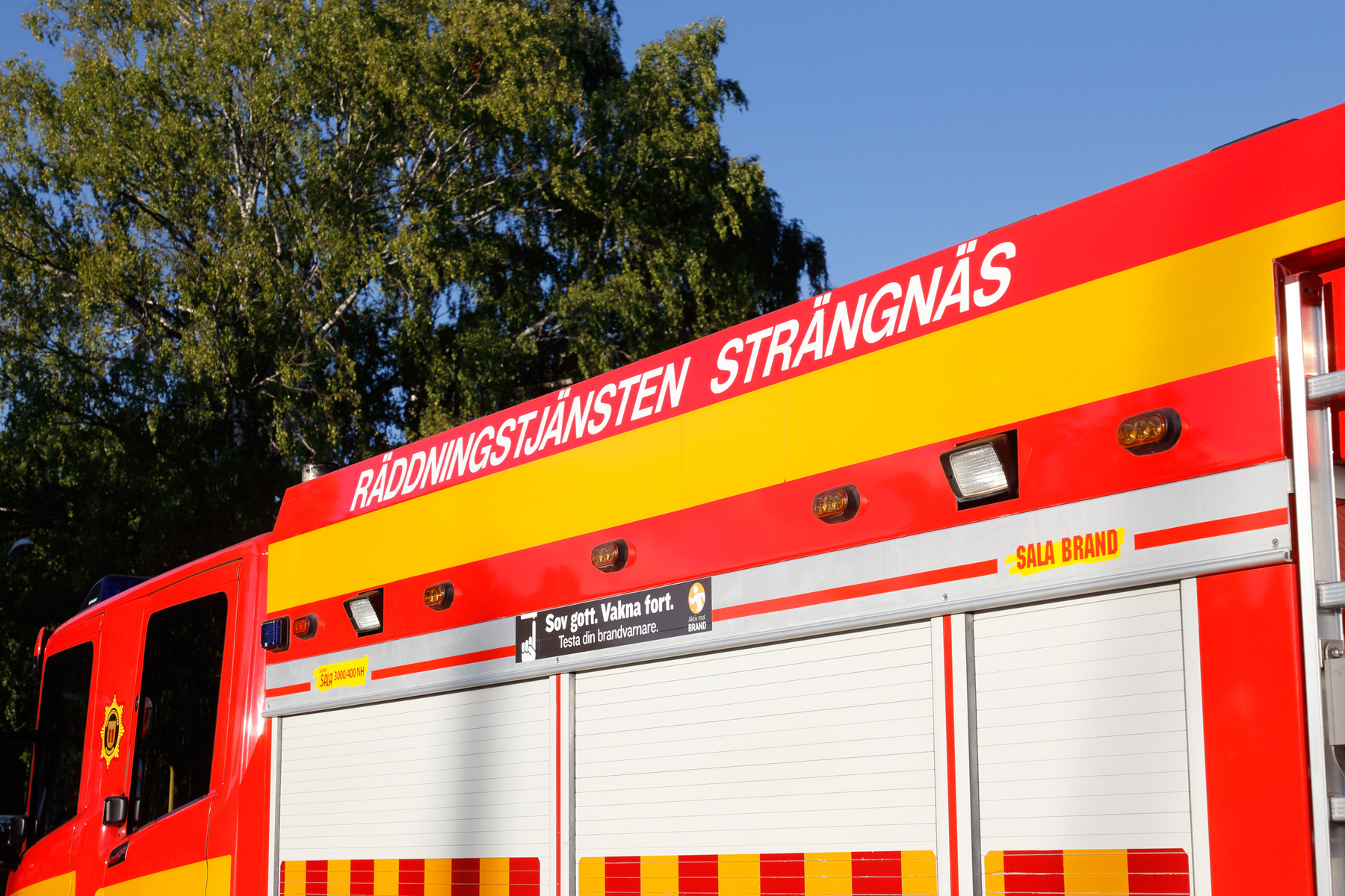 En närbild av en röd och gul brandbil utomhus under dagtid. På brandbilen står texten "RÄDDNINGSTJÄNSTEN STRÄNGNÄS" och "SALA BRAND". Ett träd med gröna löv syns i bakgrunden.