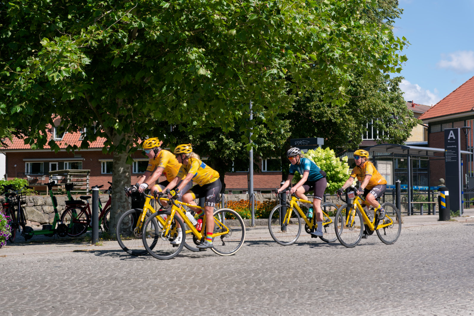 Fyra cyklister syns cyklande på en stenlagd yta, med träd och byggnader i bakgrunden. Tre av cyklisterna bär gula kläder och cykelhjälmar, medan en bär en turkos tröja och svarta shorts. Alla cyklar på gula cyklar under en solig dag.