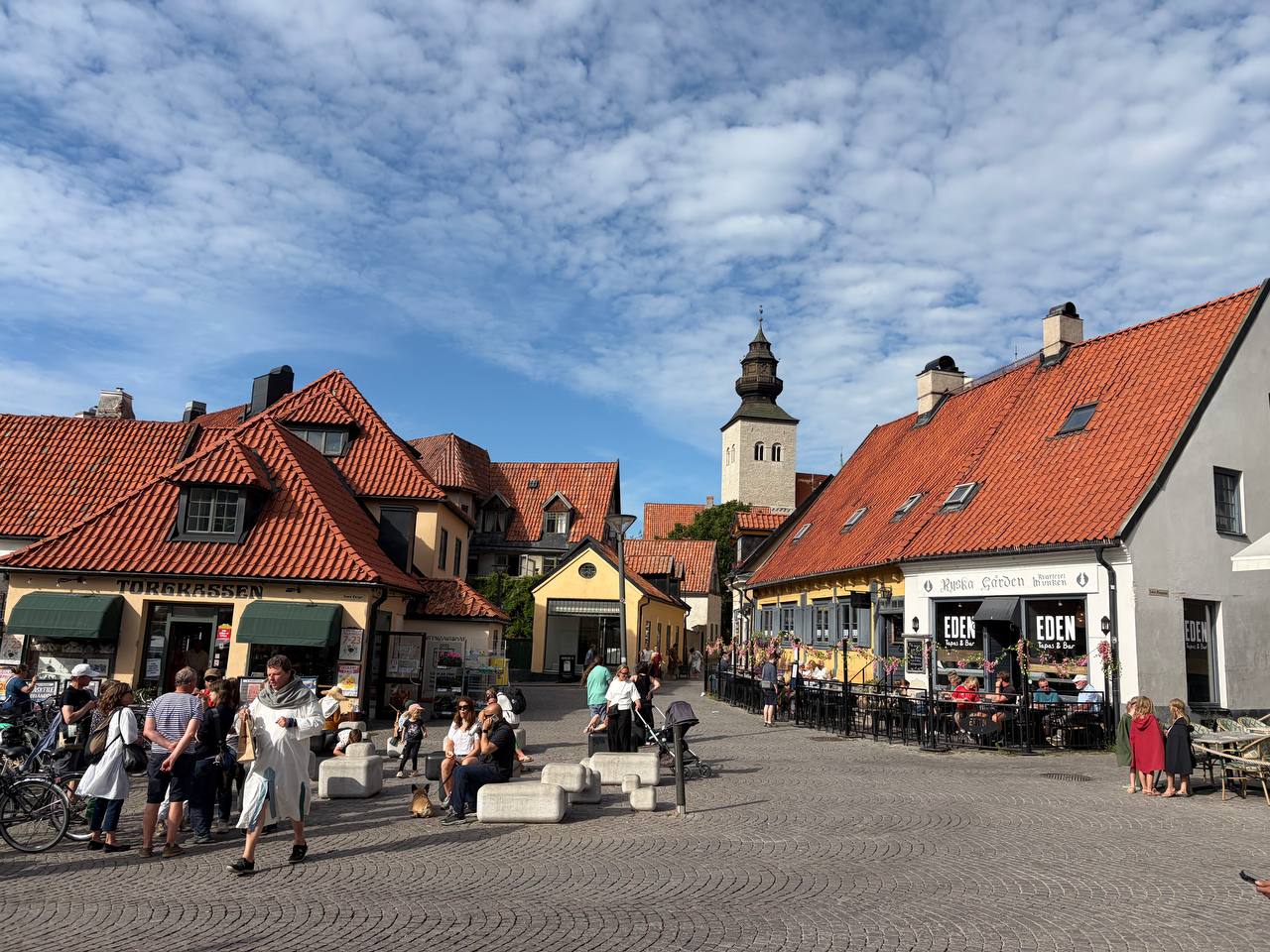 En bild av ett torg eller en öppen plats i en stad, med flera byggnader med röda tegeltak och en kyrktorn i bakgrunden. Människor promenerar eller sitter på bänkar och vid uteserveringar under en molnig himmel.