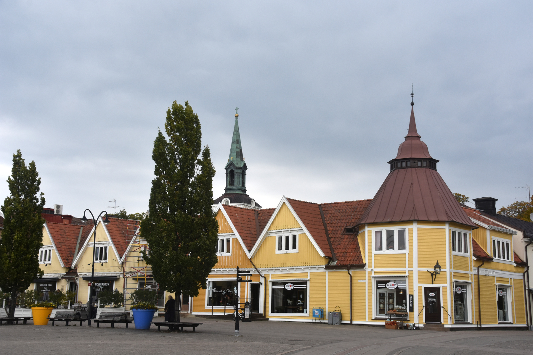 En bild som visar en torgplats framför en rad gula hus med röda tak. I bakgrunden syns ett kyrktorn med grön spira. På torget finns träd, bänkar och stora krukor i blått och gult. Himmelen är grå och molnig.