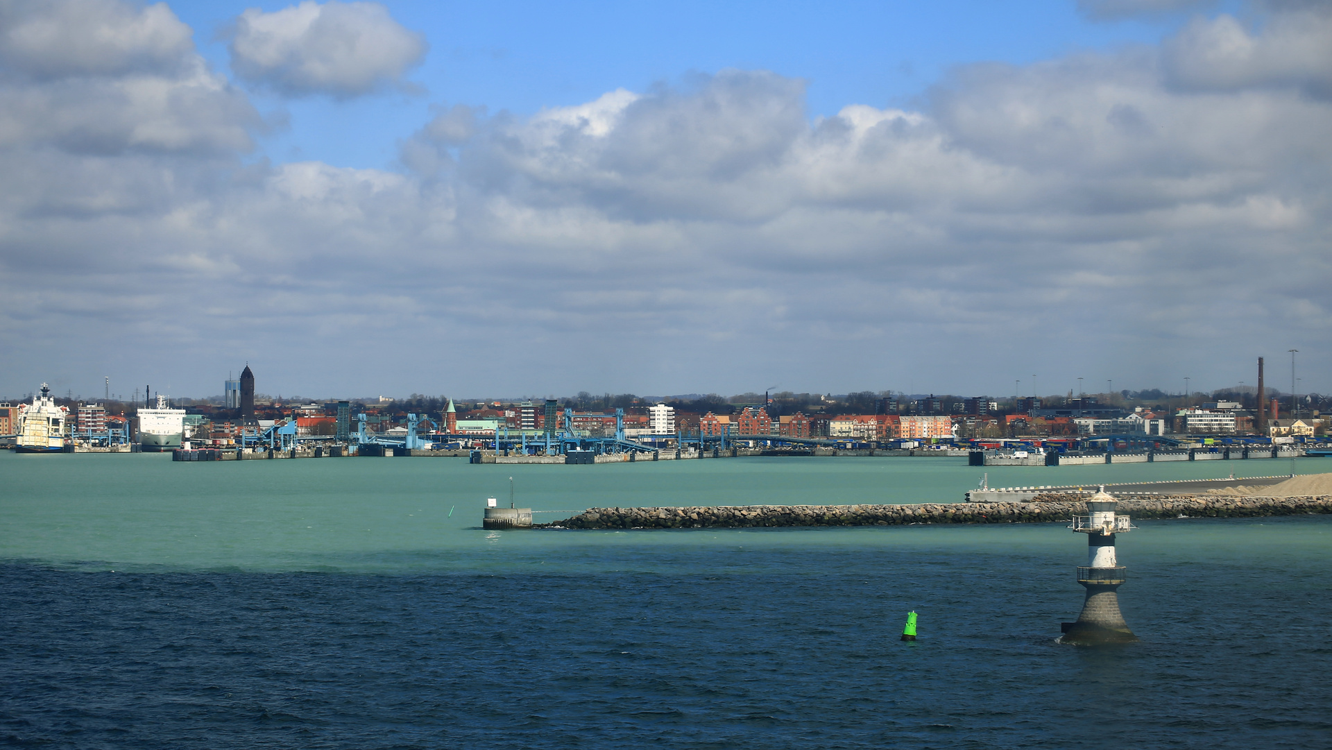 En vidvinkelbild visar en hamn och en stad under en molnig himmel. Förgrunden består av mörkblått vatten med ett fyrtorn och en grön boj. Mellan- och bakgrunden visar en hamn med flera fartyg, lastkajer och stadssiluett med byggnader i olika färger o