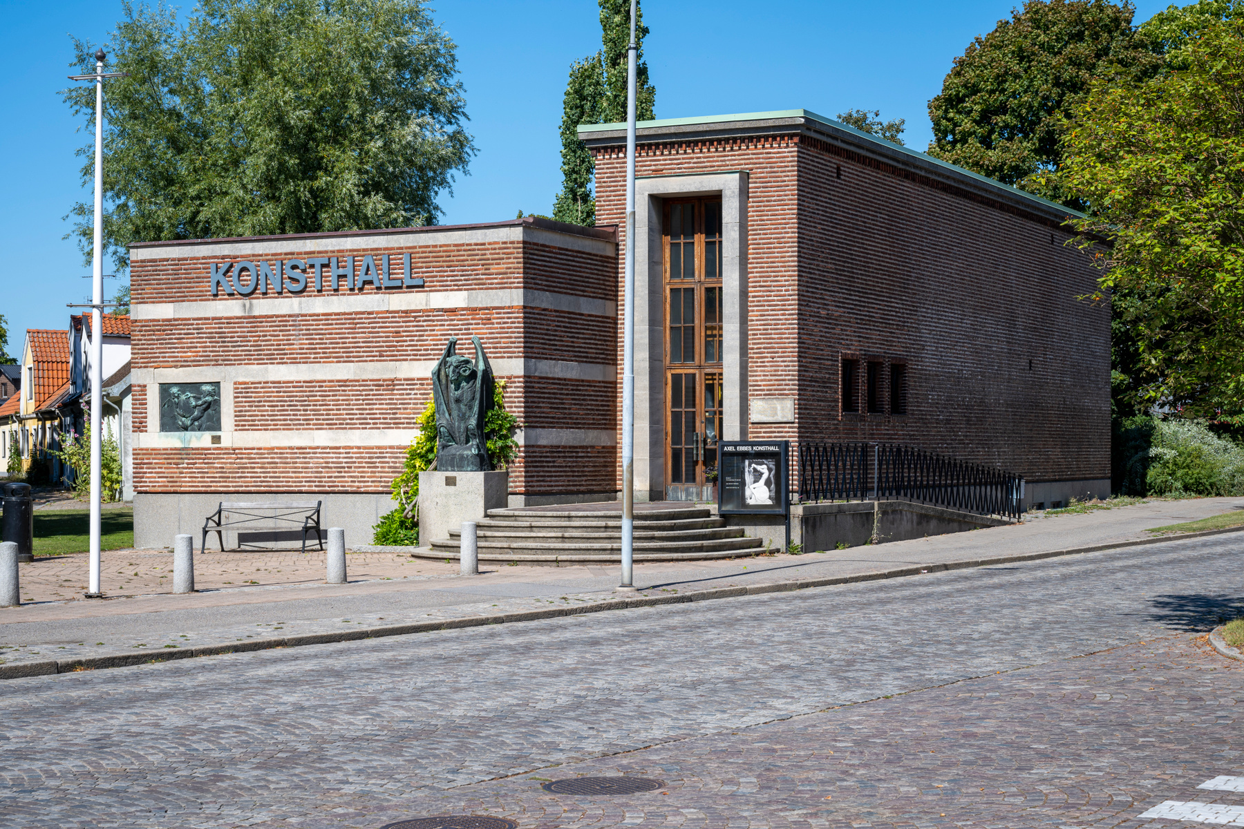 En konsthall av rött tegel med texten "KONSTHALL" på fasaden, omgiven av grönska och en kullerstensgata under klarblå himmel. Framför byggnaden syns en staty och en bänk.