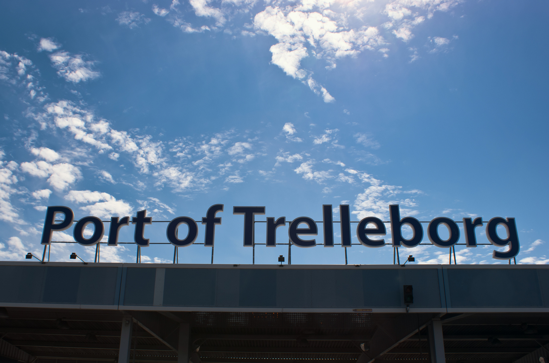 En bild tagen underifrån visar en stor skylt med texten "Port of Trelleborg" i blå bokstäver. Bakom skylten syns en klarblå himmel med vita moln och solsken. En byggnad eller konstruktion av något slag syns delvis under skylten.