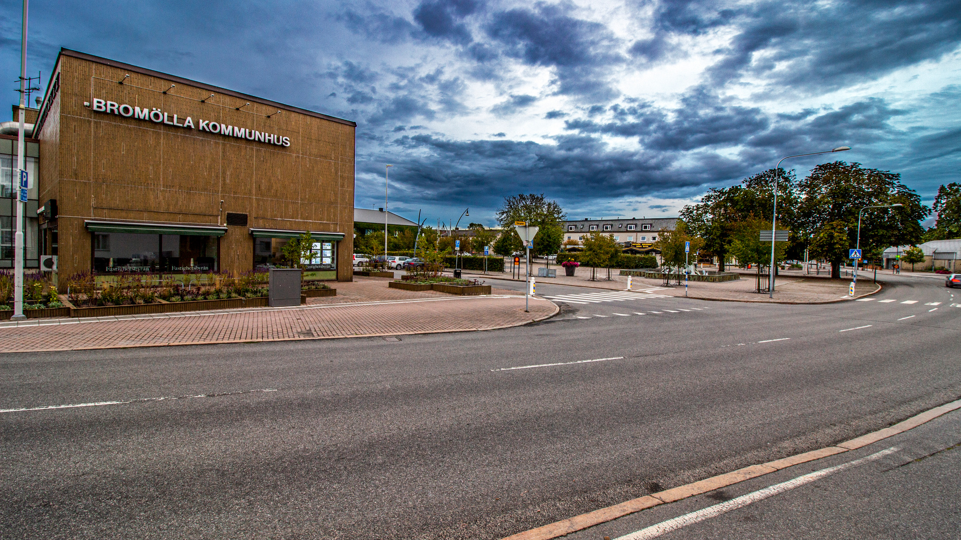 En bild som visar Bromölla Kommunhus på vänster sida, med en väg i förgrunden och ett torg med träd och en övergångsställe till höger. Himlen är molnig och grå.