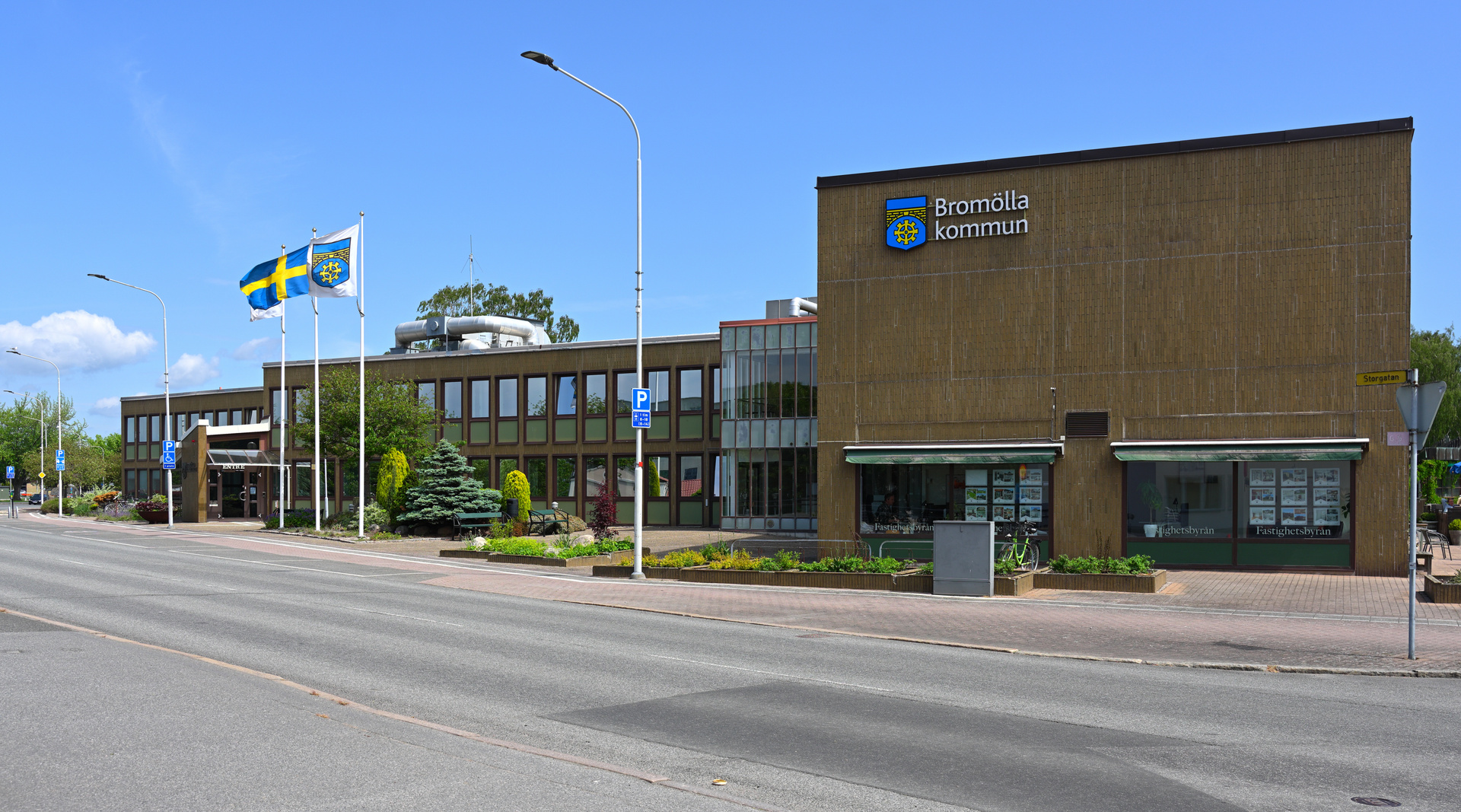 En bild som visar Bromölla Kommuns byggnad med svenska och kommunala flaggor i förgrunden, bredvid en väg. På byggnaden syns skyltar med "Bromölla kommun" och "Fastighetsbyrån". En parkeringsskylt och en skylt med "Storgatan" syns också.