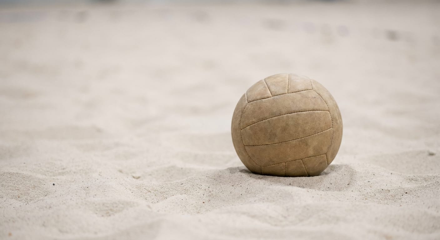 En beige volleyboll med sömmar ligger på ljus sand. Sanden har små mörka partiklar och ojämn yta. Volleybollen är placerad i bildens högra del och bakgrunden är oskarp.