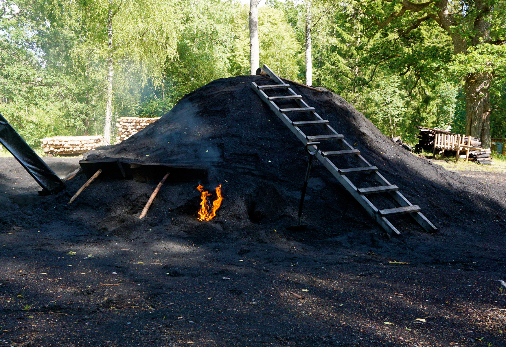 En stor, mörk kulle, sannolikt en kolmila, står utomhus med en eld synlig vid dess bas. En trästege lutar mot kullen. I bakgrunden syns gröna träd och staplar av ved. Markytan framför kullen är också mörk.