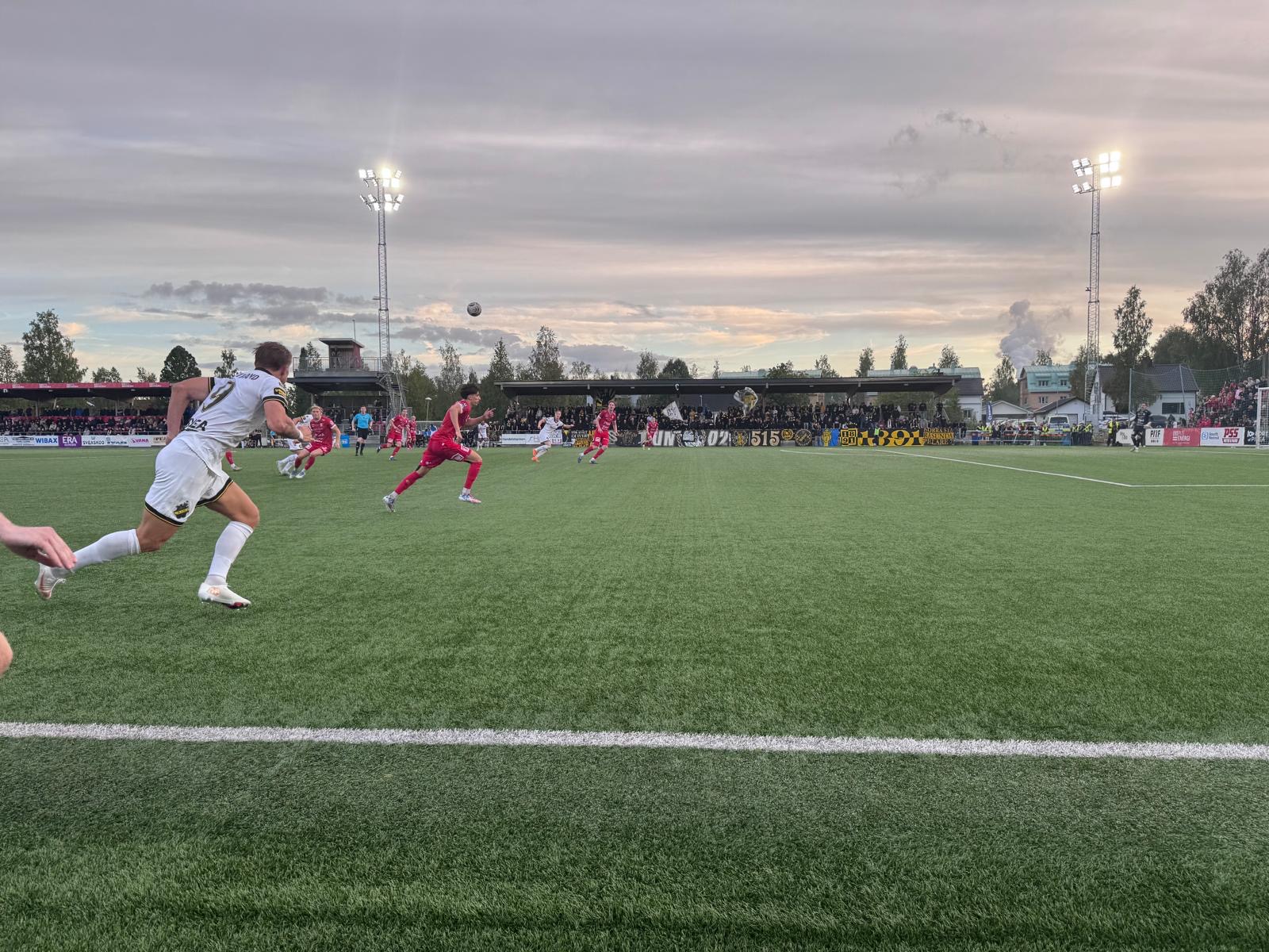 En fotbollsmatch pågår på en grön konstgräsplan med flera spelare i röda och vita/svarta dräkter. En fotboll syns i luften. I bakgrunden syns åskådare på en läktare, träd och byggnader. Två höga belysningsmaster lyser upp planen under en molnig himme