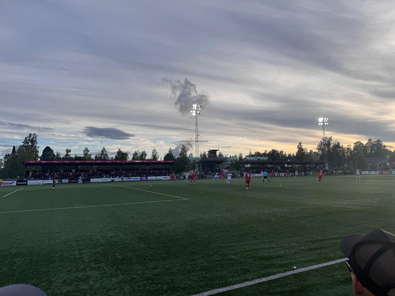 En bild visar en fotbollsmatch på en grön konstgräsplan under en molnig himmel. Två lag i vita respektive röda tröjor spelar på planen, som omges av läktare med publik och träd. Två strålkastartorn med tända lampor syns, och solen står lågt vid horis