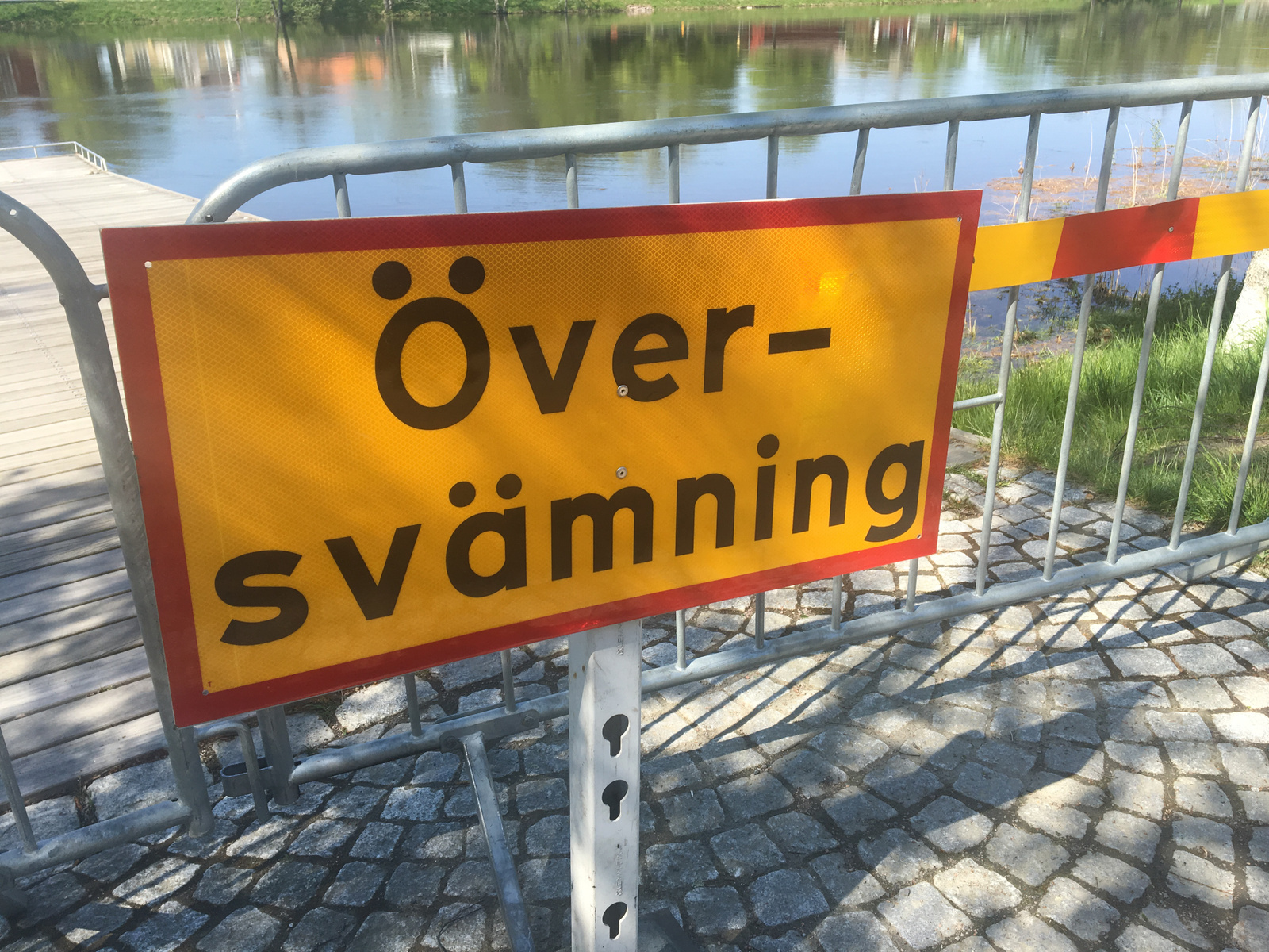 En gul varningsskylt med texten "Översvämning" i svart är uppsatt på ett grått metallstaket i förgrunden. Bakom staketet syns en vattenyta som reflekterar ljus och hus. I förgrunden syns också mark med gatsten och gräs.