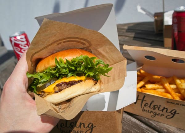 En hand håller en cheeseburgare inslagen i brunt papper, med sallad och smält ost synliga. I bakgrunden syns en öppen kartong med pommes frites och logon 'Helsing burger' samt texten 'SMAKLIG MÅ'. En röd läskburk med 'Coca-Cola' tryckt på syns snett