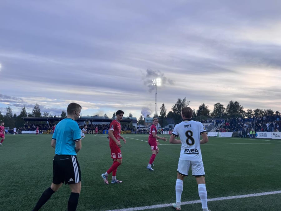 En bild tagen underifrån på en fotbollsplan där flera spelare och en domare är synliga. Domaren i turkos tröja står med ryggen mot kameran, och framför honom finns två spelare i röda dräkter. En fjärde spelare i vit tröja med nummer 8 och texten "HOV
