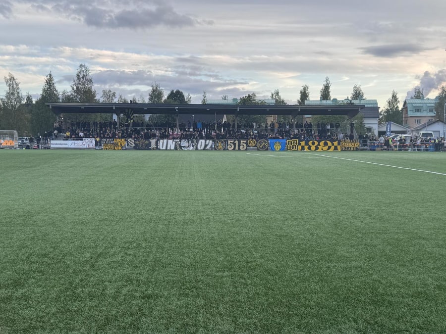 En bild visar en fotbollsplan med konstgräs och en läktare i bakgrunden. På läktaren sitter och står människor, och flera banderoller med text och logotyper är uppsatta framför publiken. I bakgrunden syns träd och byggnader under en molnig himmel.