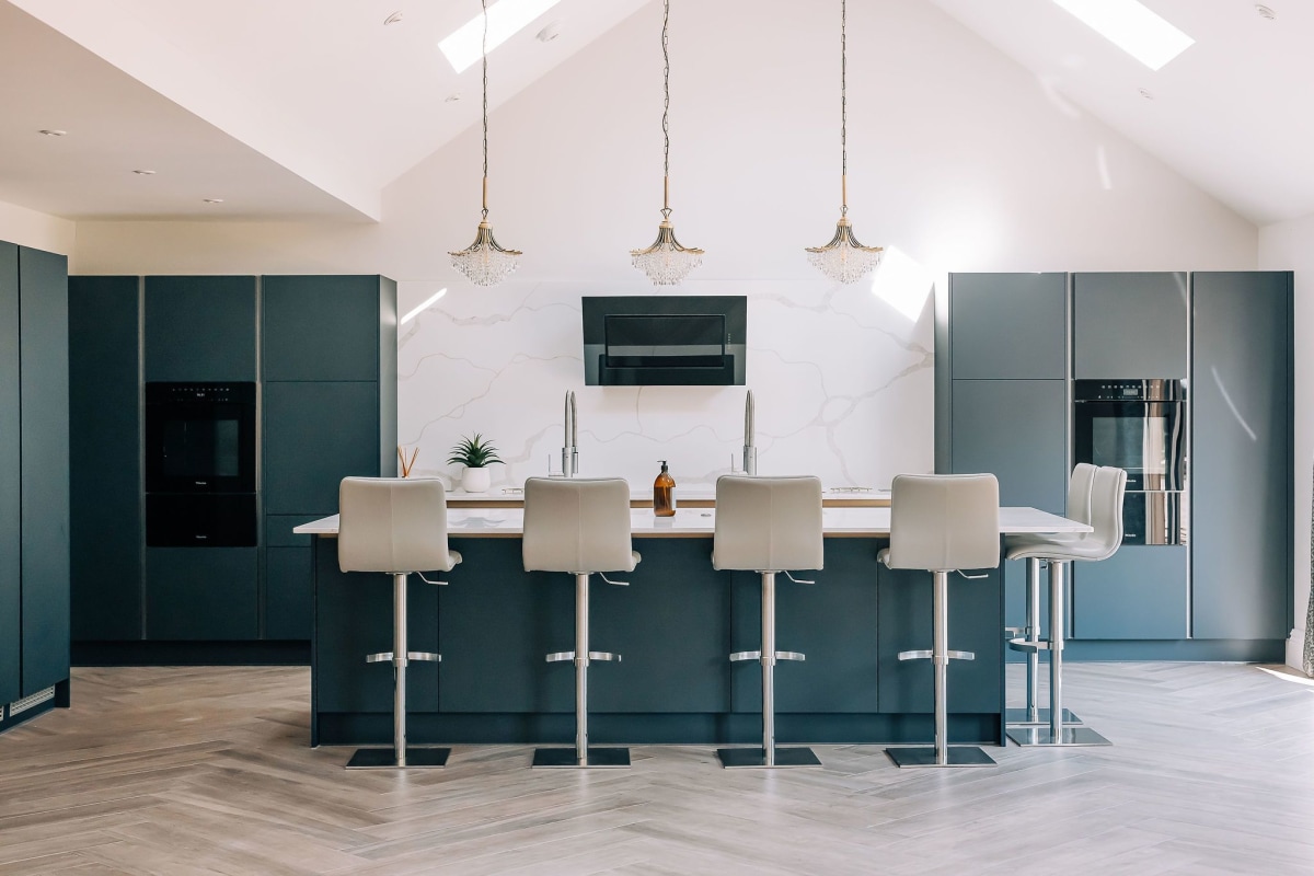 A rich blue modern kitchen with wooden herringbone floor and cream bar stools, with white quartz worktop