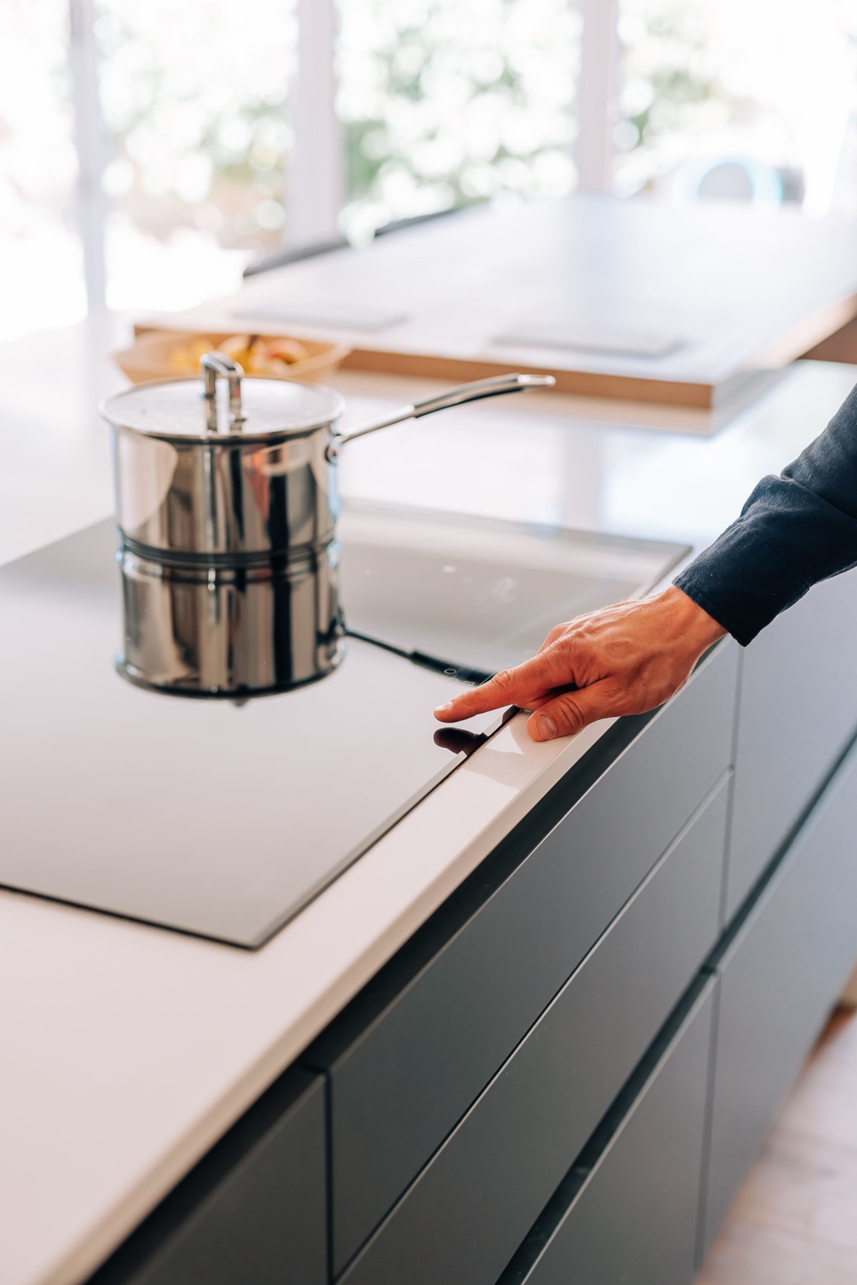 A hand turning on an induction hob on a kitchen island with blue handleless units beneath