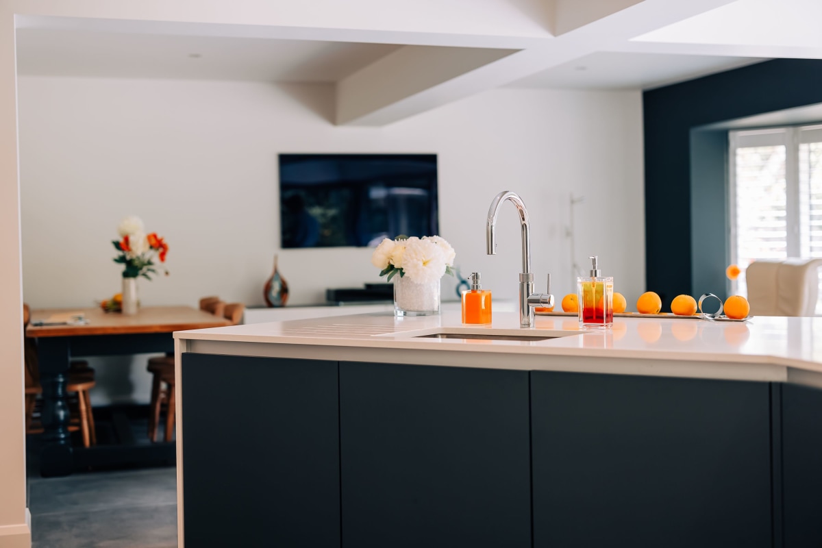 A dark blue contemporary kitchen island with white quartz worktop and a living room with television in the background