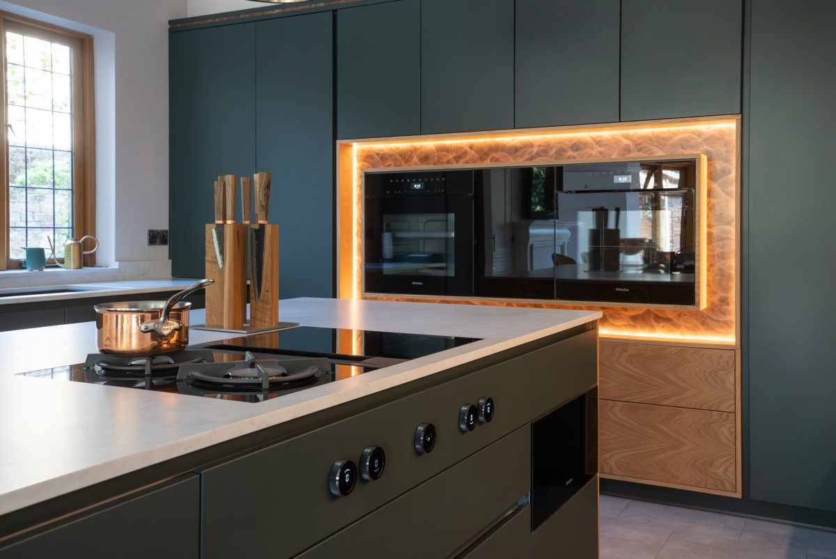 A kitchen with dark cupboards in the background and illuminated oven surround, with an island in the foreground with white matt quartz worktops