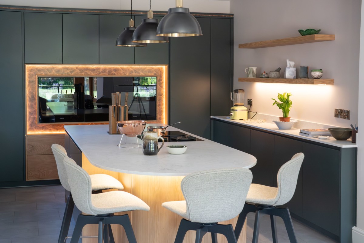 A kitchen island with white stools, with dark green cabinetry and ovens in the background