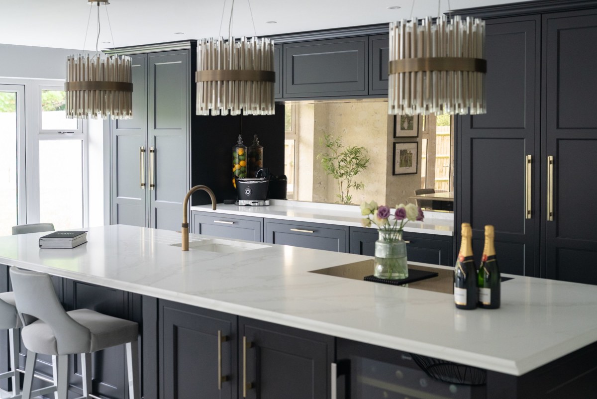 A black shaker kitchen with brass handles and a white worktop, with three pendant lights hanging above