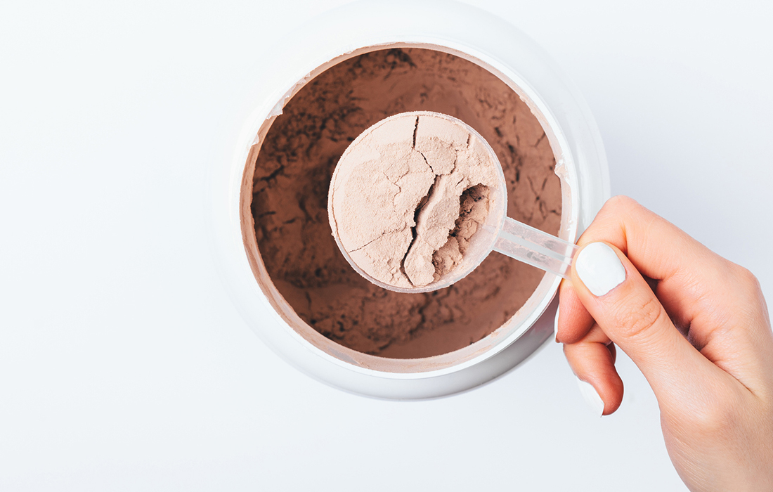 Hand holding a scoop of protein powder over a tub