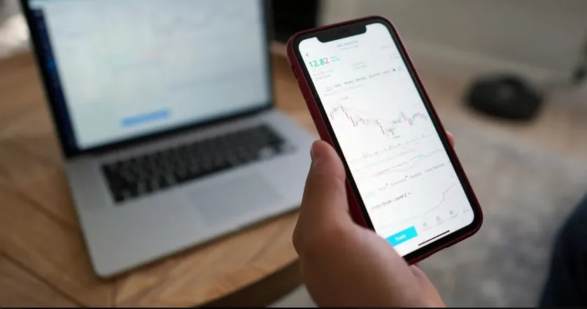 Hand holding smartphone displaying stock chart on copy trading platform with laptop in background.