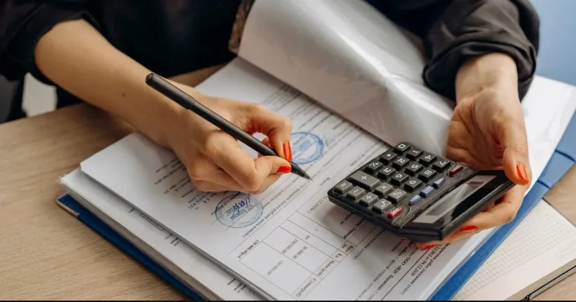 Hands holding pen and calculator reviewing forex trading documents and regulatory compliance paperwork.