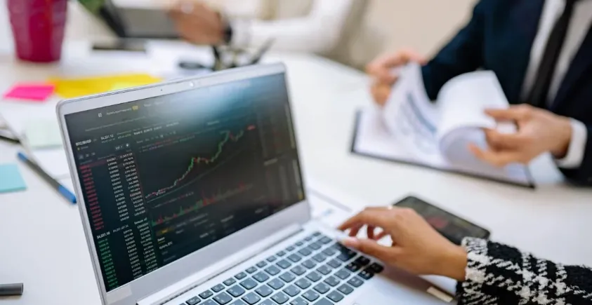 Trader analyzing stock charts on laptop with colleagues reviewing financial data in background.