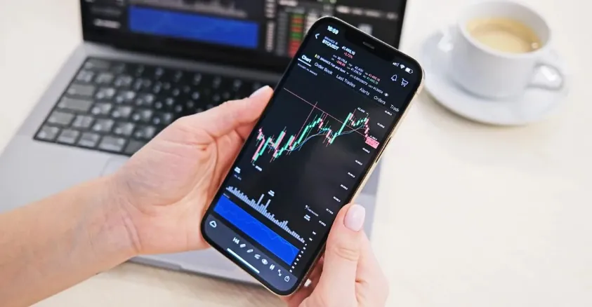 Hand holding smartphone displaying live stock charts with trading app interface and laptop in background.