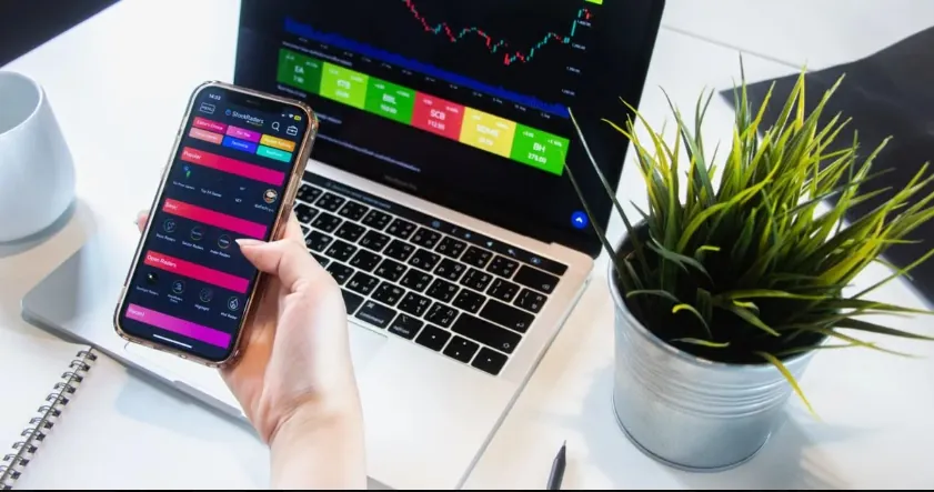Hand holding smartphone displaying stock market data beside laptop with trading charts and green plant on desk.